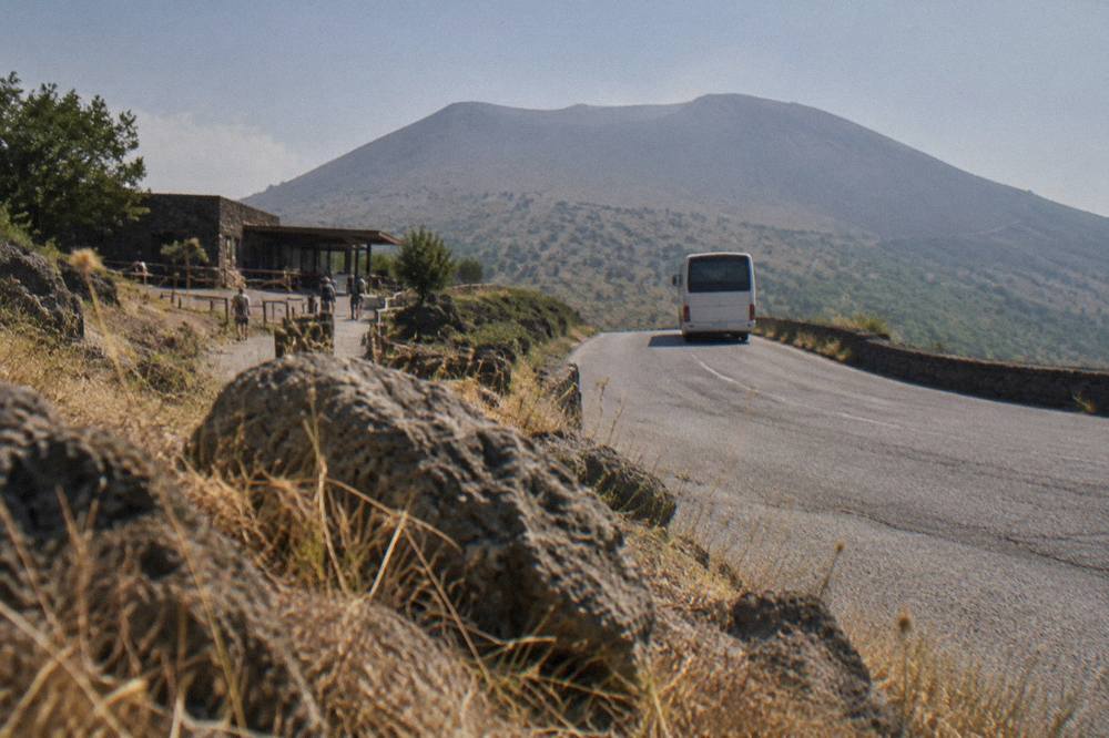 A regional bus at a stop on the lower slopes of the volcano with dry grass and lava stone beside the road