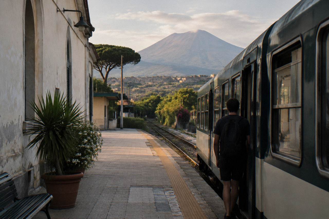 A narrow-gauge regional train at a station platform under a summer sky near Naples