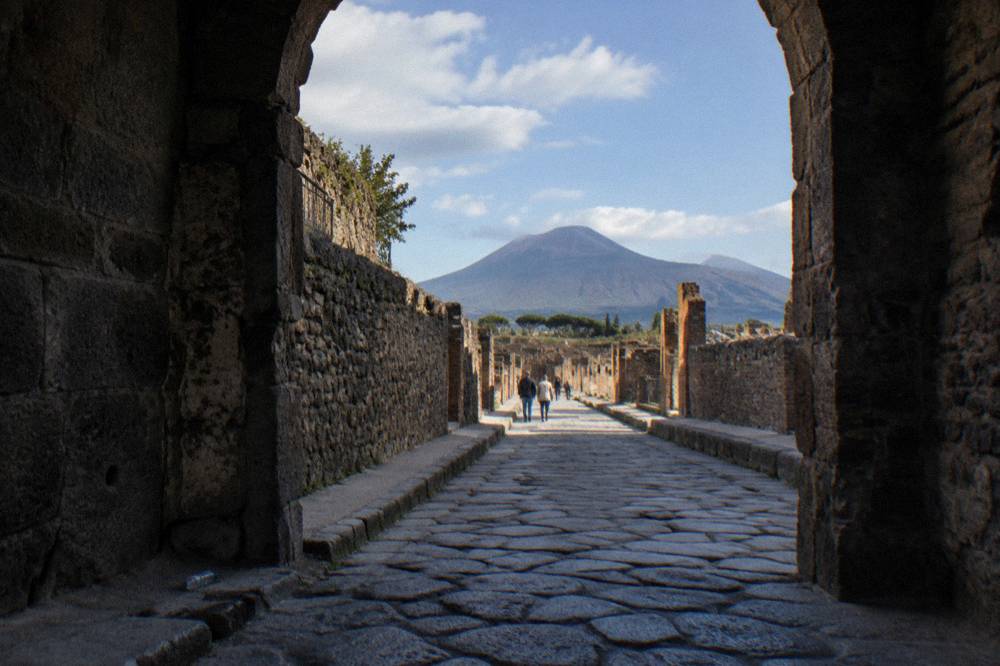 Stone-paved street in the Pompeii ruins with Vesuvius rising in the background under scattered clouds