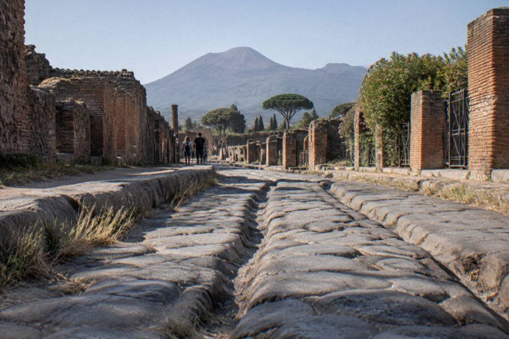A stone-paved ancient street in Pompeii with worn cart ruts and ruined walls under a clear sky
