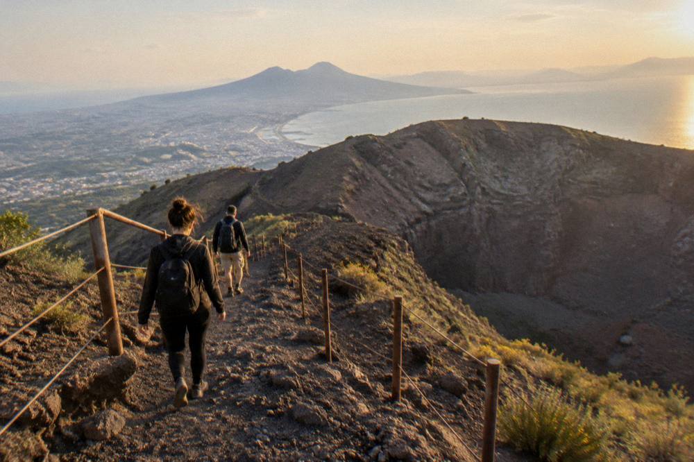 Hikers descending the gravel crater path in late afternoon light with the Bay of Naples below