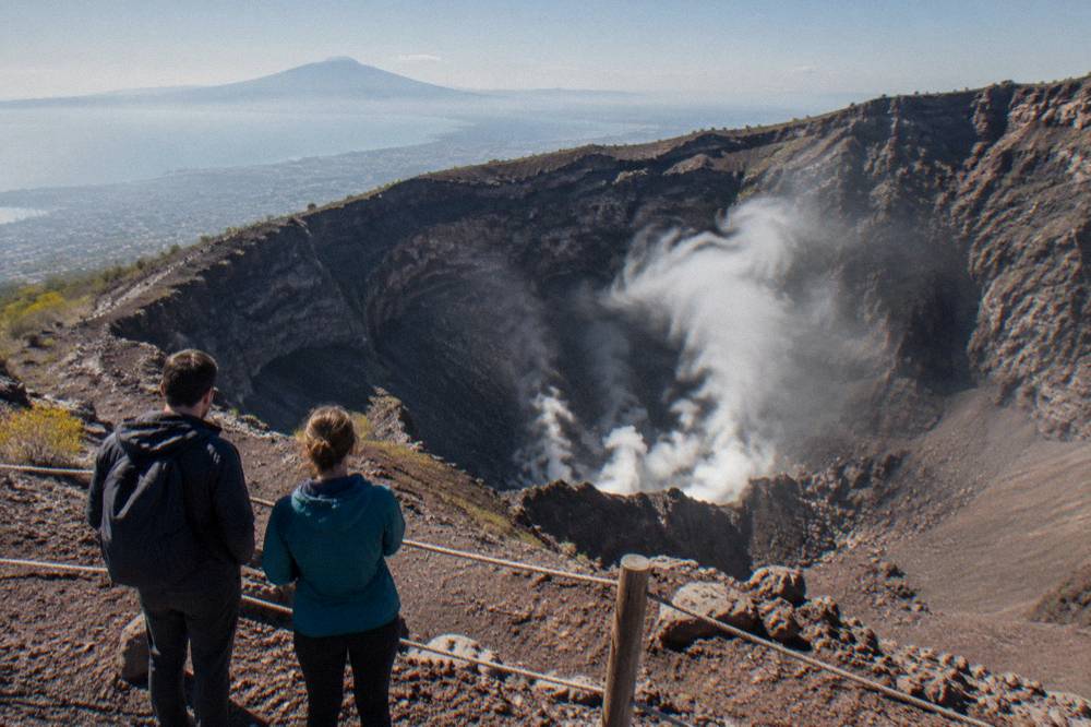 Hikers in windbreakers standing at the crater rim viewpoint above steaming fumaroles