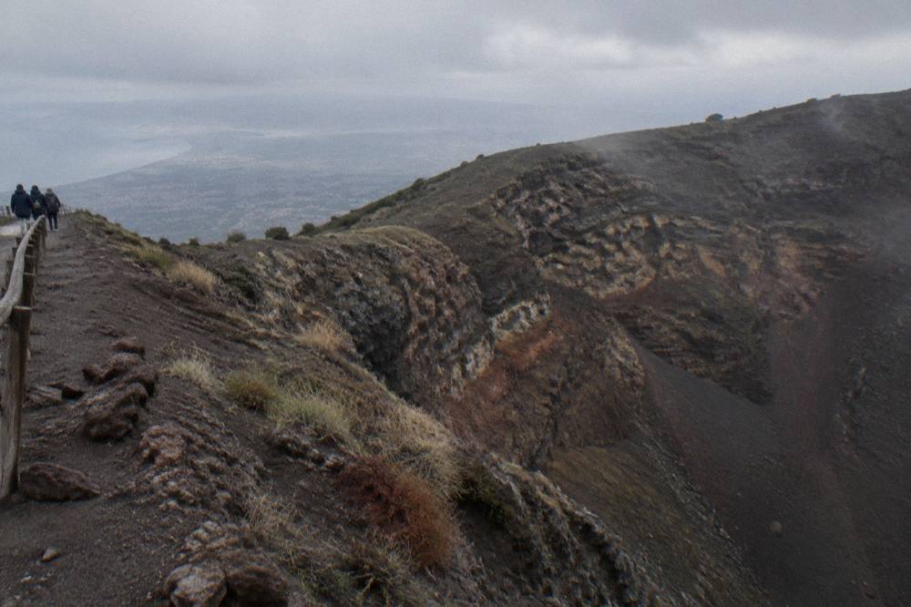 The rim of the Vesuvius crater with layered dark rock walls and a hazy plain beyond.
