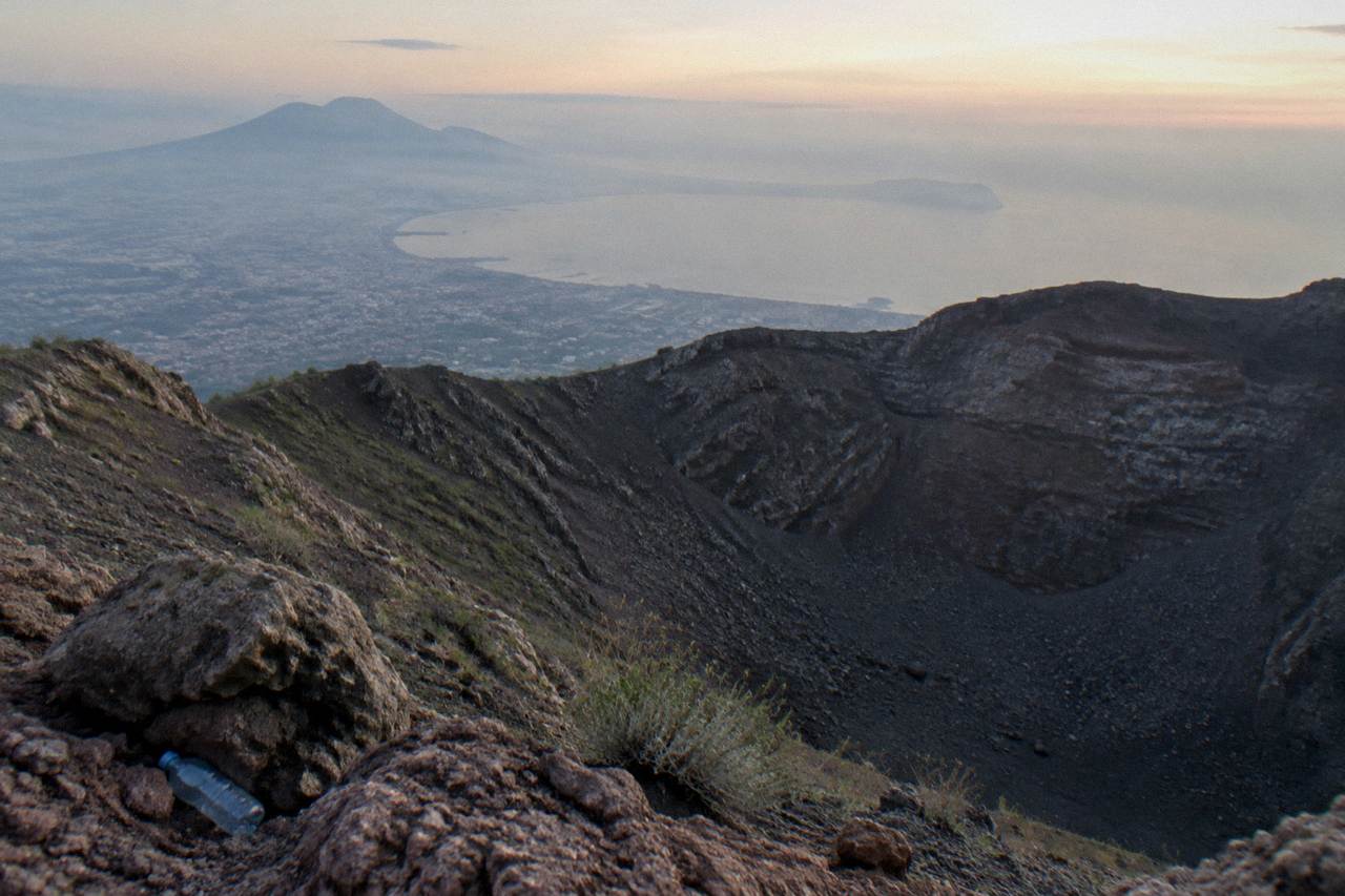 The crater of Vesuvius under a pale sky, dark volcanic rock slopes descending toward the Bay of Naples.