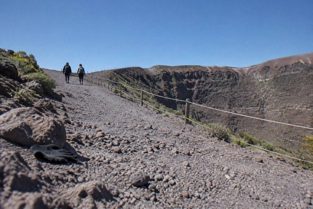 Hikers on a wide volcanic gravel path approaching the Vesuvius crater rim under clear sky