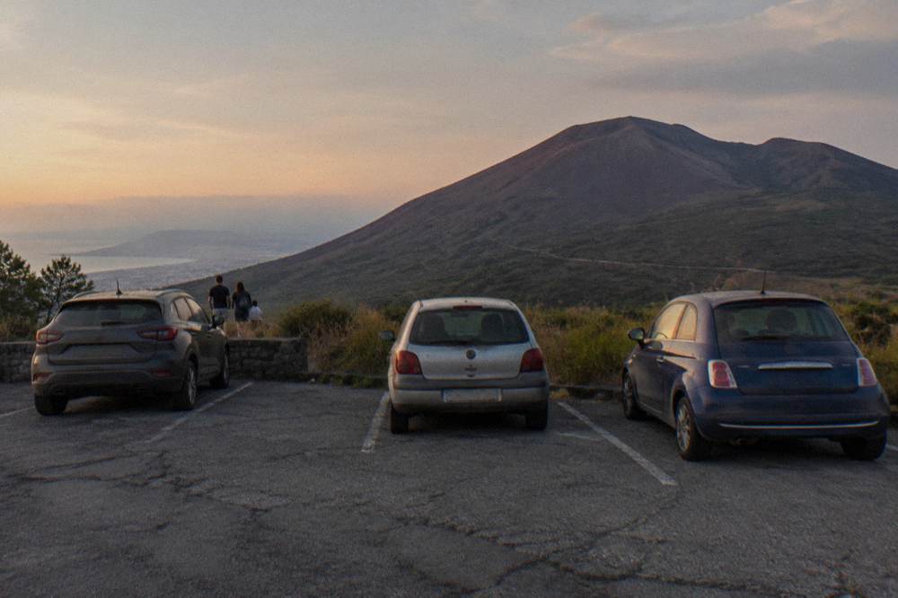 A winding mountain road above Ercolano with parked cars and the volcano slope rising ahead