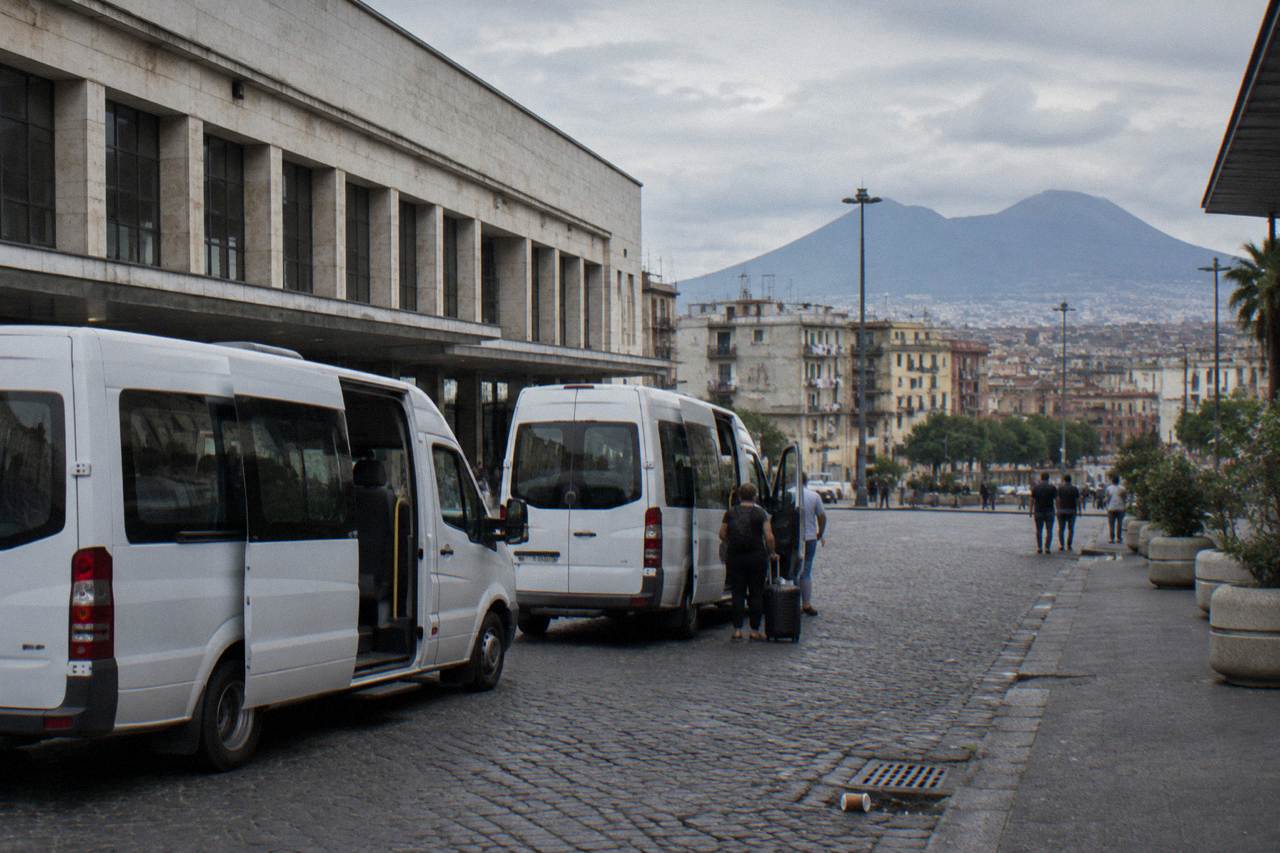 Tour minibuses parked on a street beside the Naples station square with Vesuvius rising in the distance