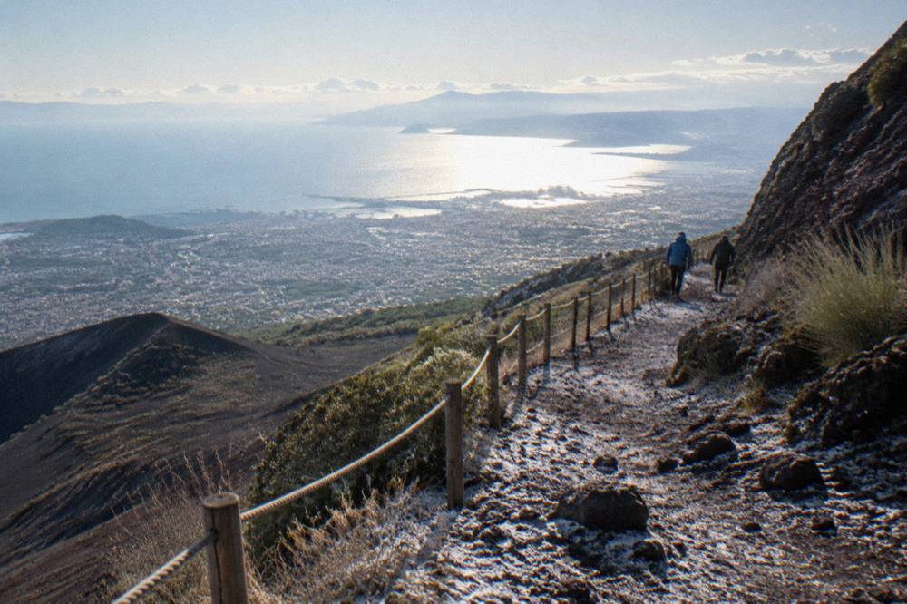 Hikers in windbreakers on the Vesuvius crater path with the bay far below in sunlight