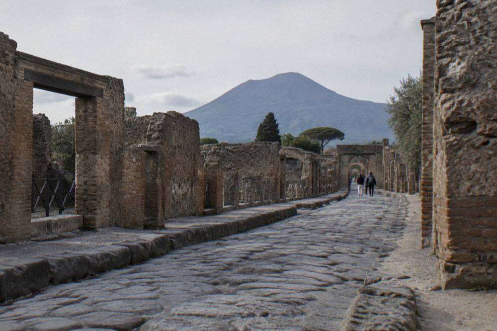 A stone path through an ancient Pompeii ruin with the profile of Mount Vesuvius rising in the background