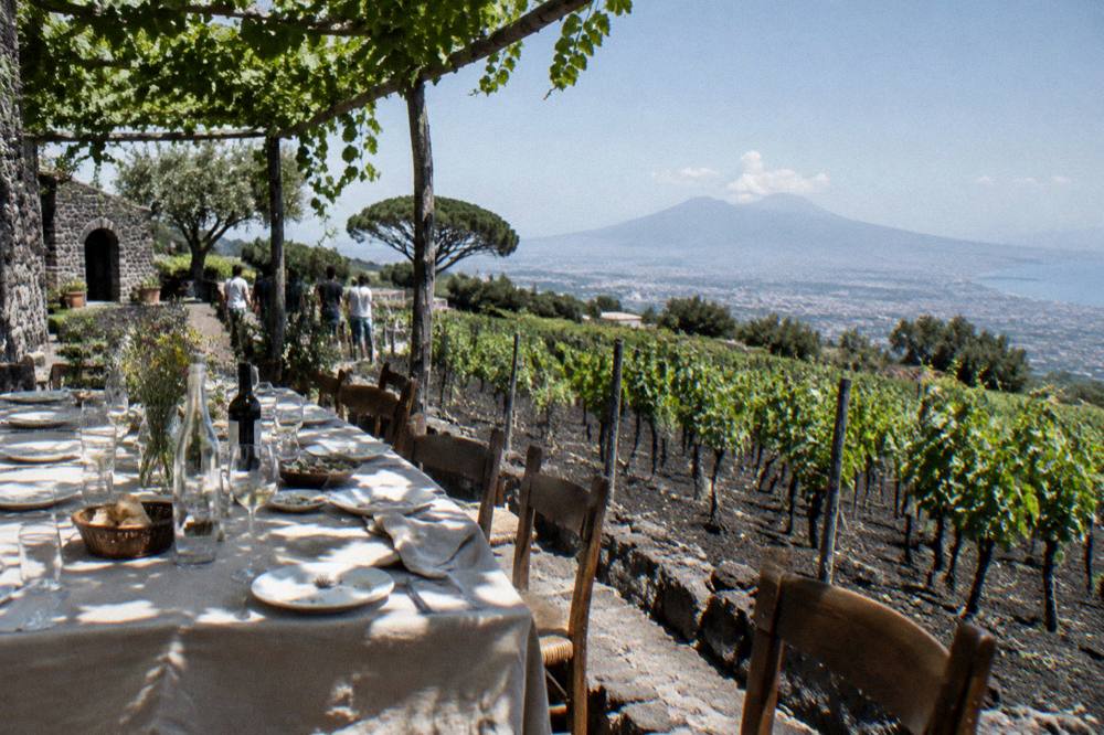 An outdoor lunch table on a winery terrace overlooking terraced vineyards on the slopes of Vesuvius