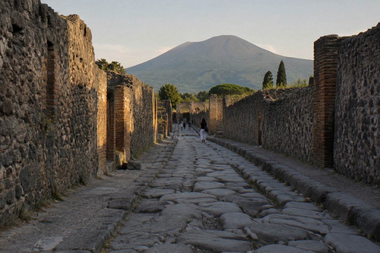 The stone streets of Pompeii with the slopes of Mount Vesuvius rising in the background under a clear sky.