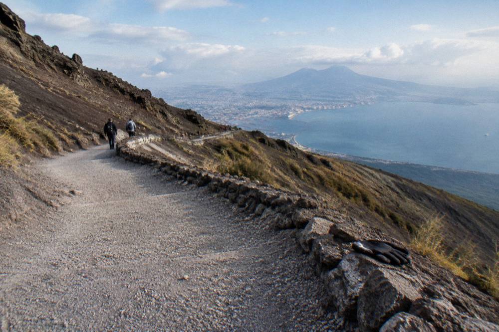 A broad gravel switchback path on a dark ash slope with the Bay of Naples in the distance