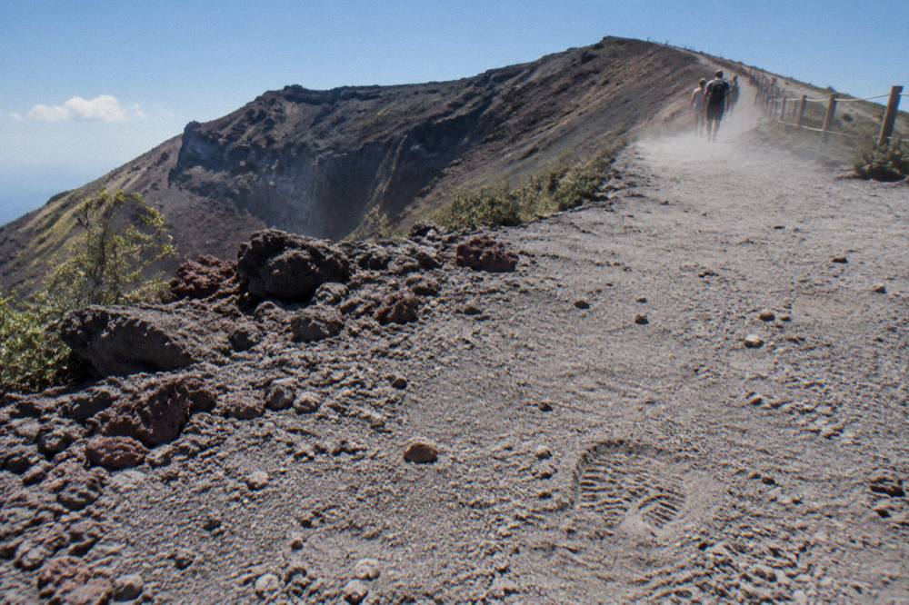 A dusty trail winding up an ash slope toward a crater rim in bright sun