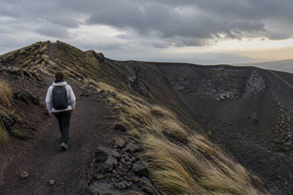 A hiker in a light jacket walking the dusty ash path toward the Vesuvius crater rim