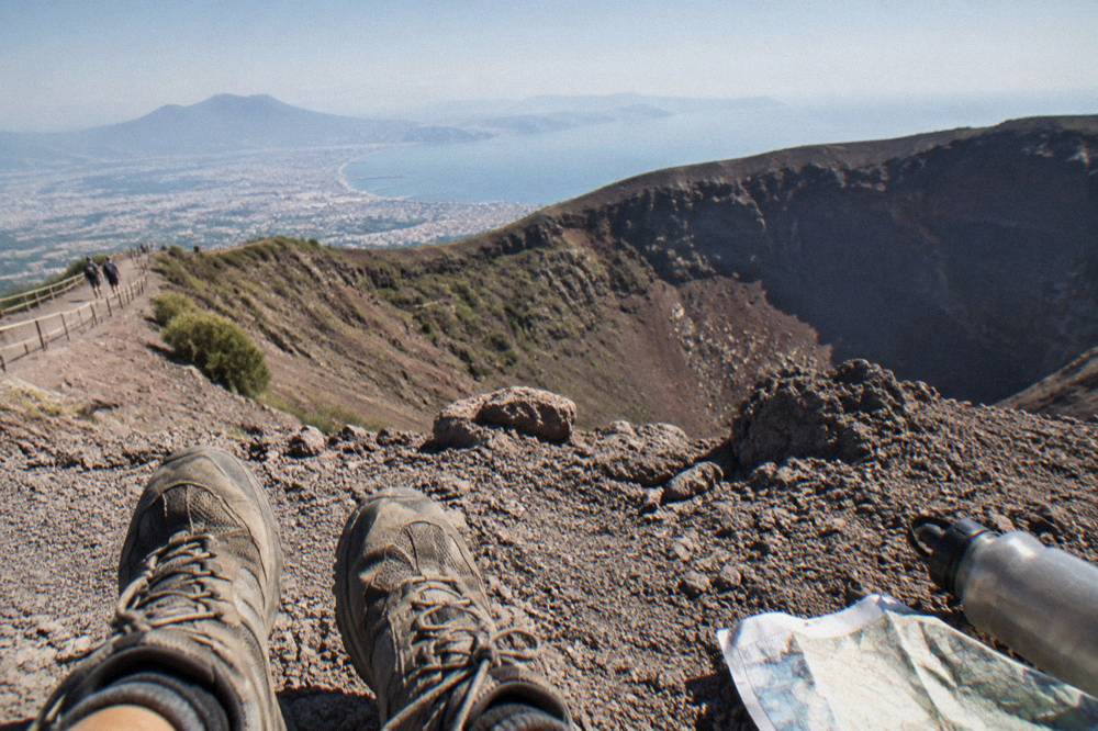 Walking shoes standing on pale volcanic ash beside the crater trail with the Bay of Naples far below