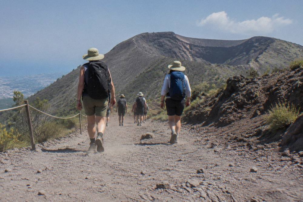 Hikers in sun hats walking the dusty gravel path toward the Vesuvius crater