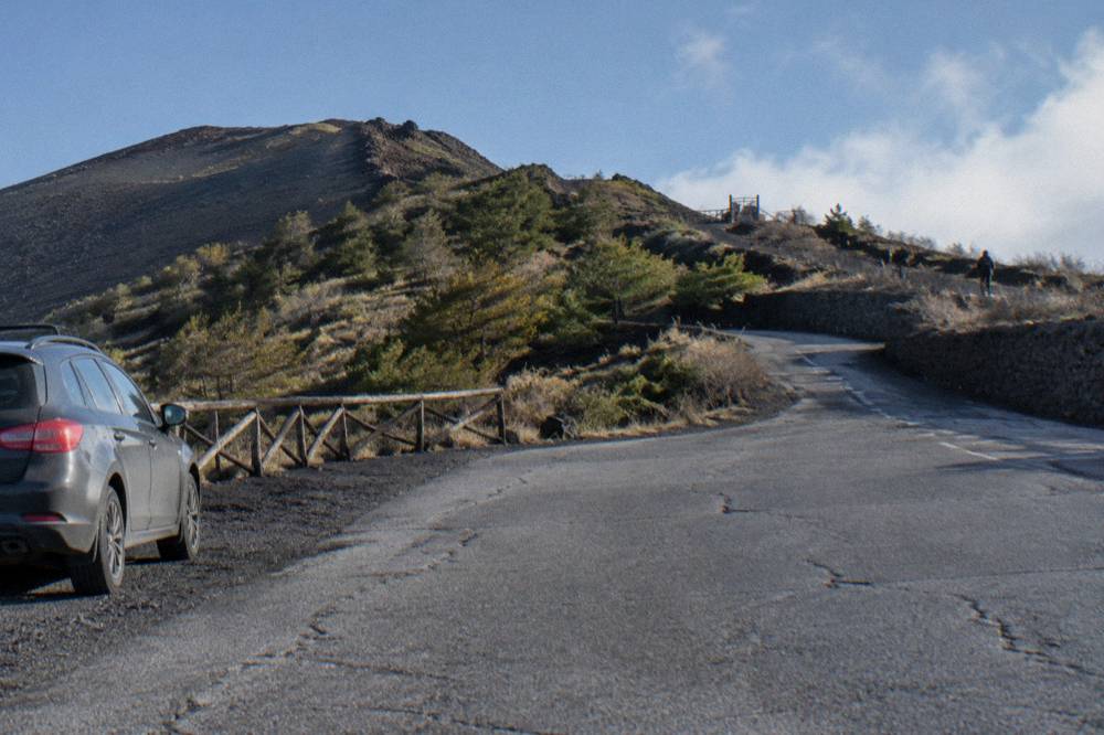 Winding mountain road with volcanic slopes and parked cars near a trailhead gate